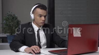 年轻成功的非洲商人穿着西装，戴着耳机在电脑上工作，在工作中玩电脑游戏